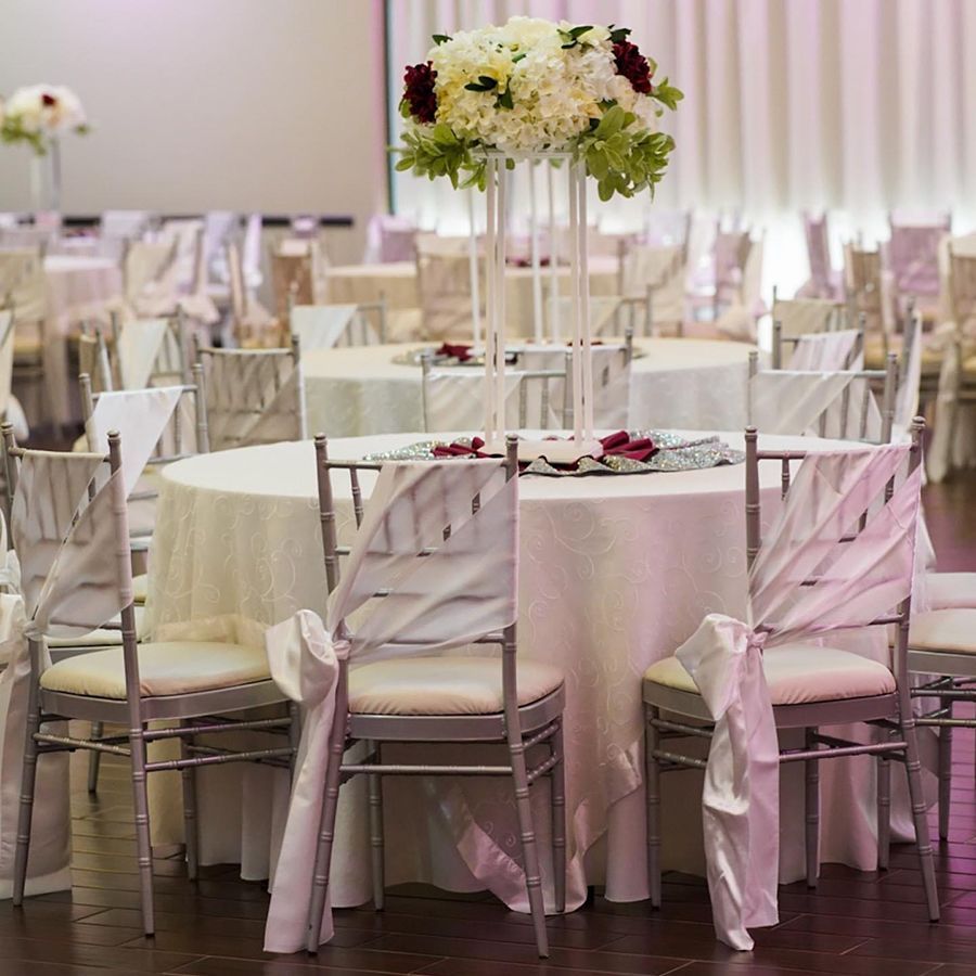 Wedding reception with round tables, floral centerpieces, and silver chairs draped with white fabric.
