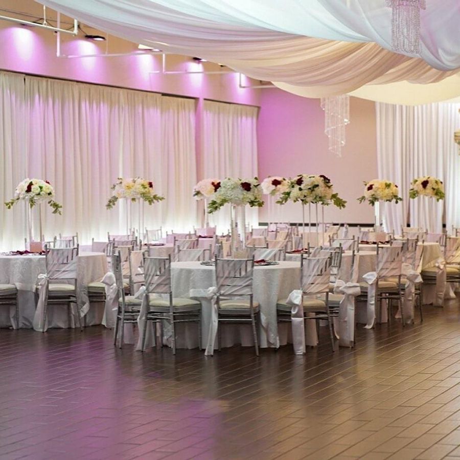 Elegant event hall set for a celebration, with tables, floral arrangements, draped fabric, and ambient lighting.