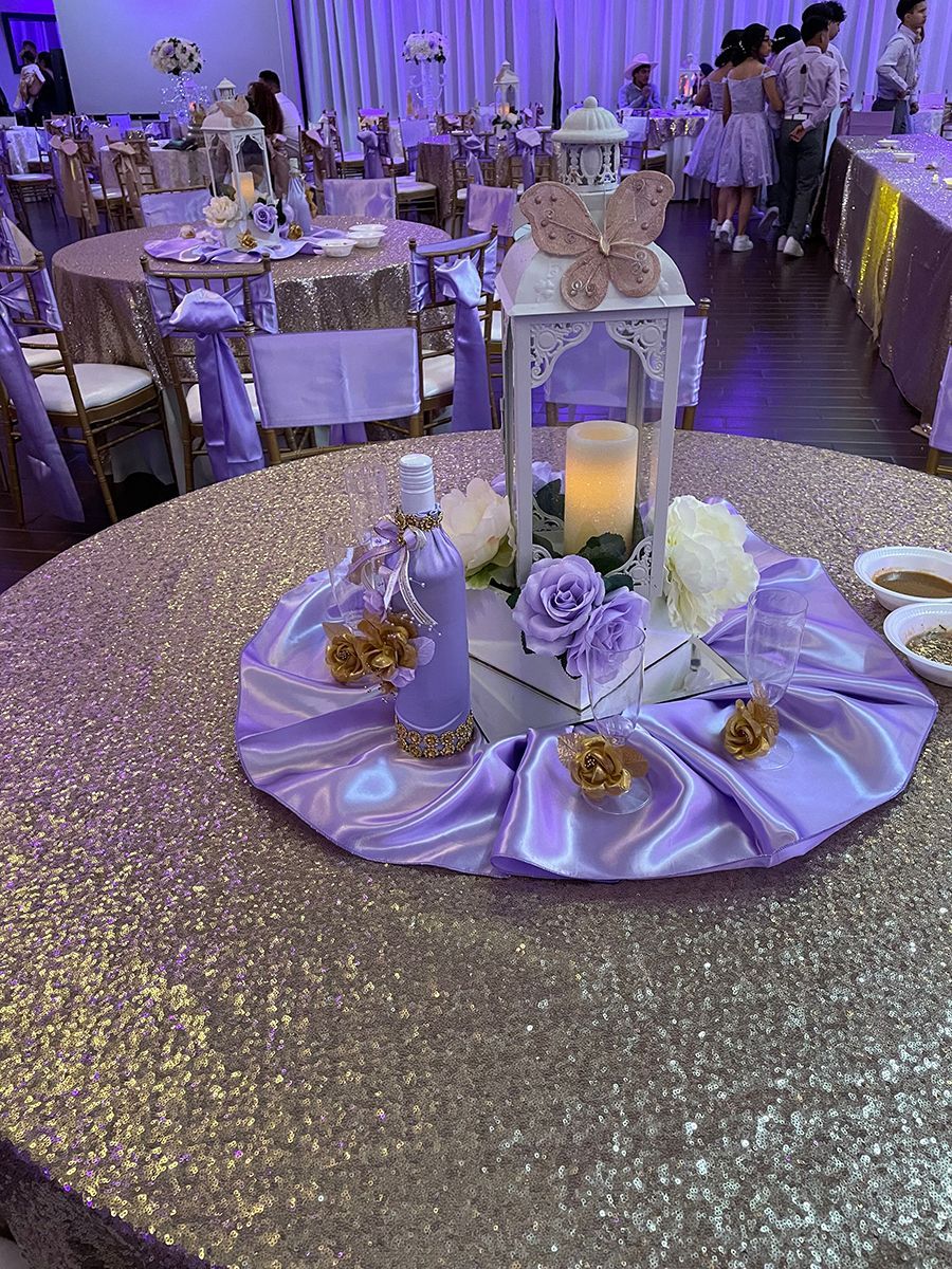 A table centerpiece with a lantern, flowers, and a bottle on a gold sequin tablecloth in a venue.