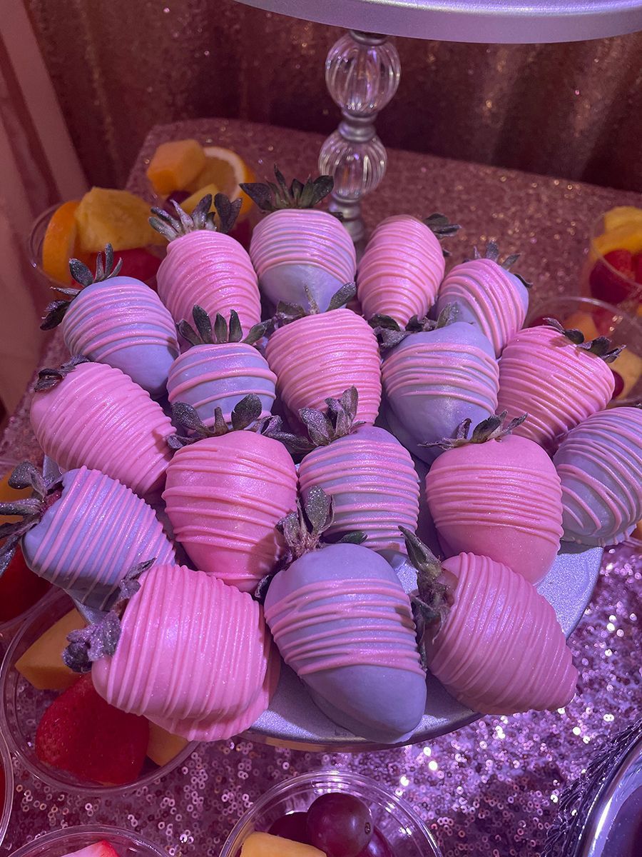 Chocolate-covered strawberries on a tiered platter, some pink and some blue, surrounded by fruit.