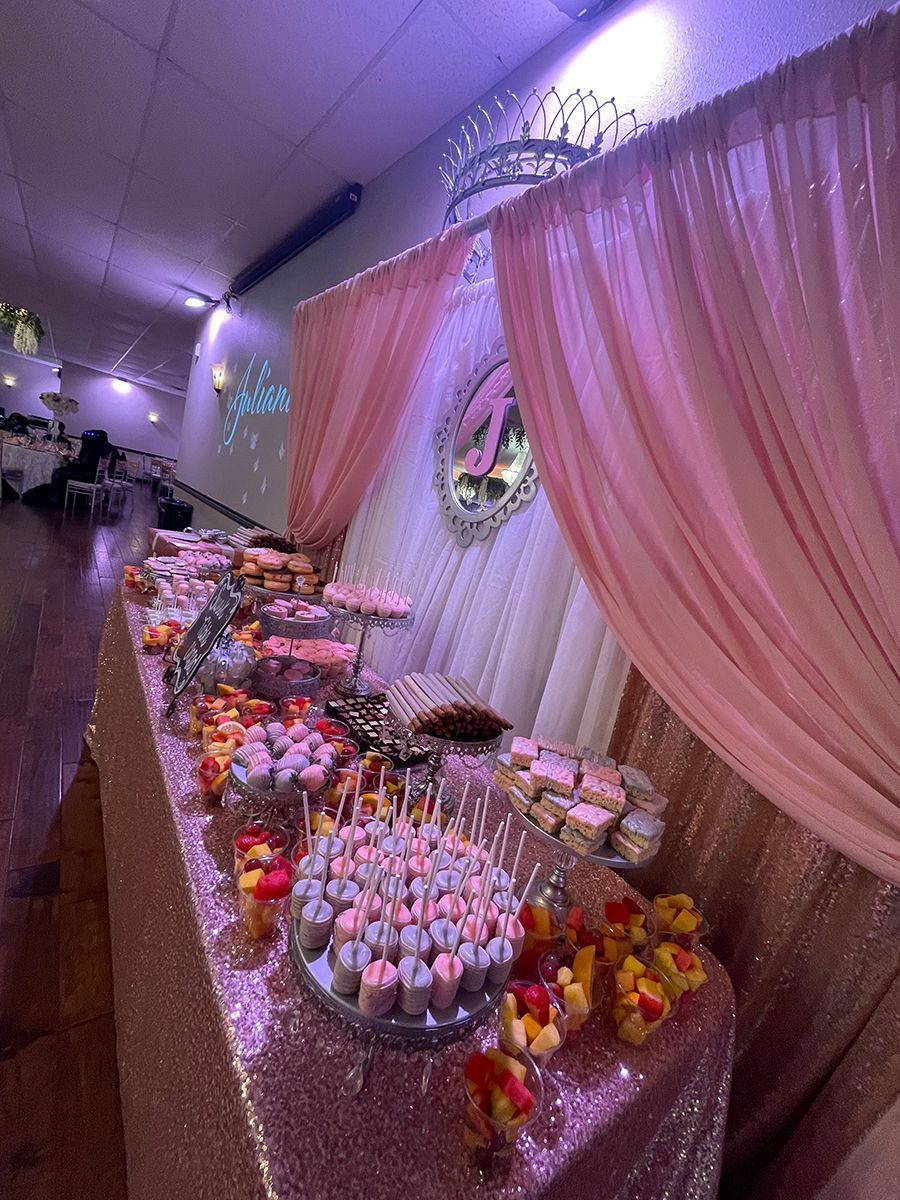 Dessert table at a party, featuring sweets on trays. Pink and gold decor with drapes and a backdrop with the letter 
