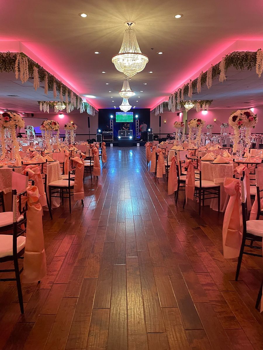 Ballroom decorated for an event, pink and gold theme, tables set, stage at the far end, chandeliers.