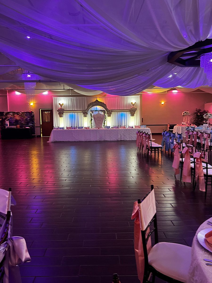 Wedding reception hall with draped ceiling, wood floor, tables and chairs, and decorative lighting.