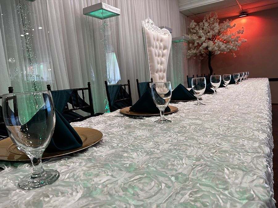 Long banquet table set with glassware, gold chargers, green napkins, and white floral tablecloth.