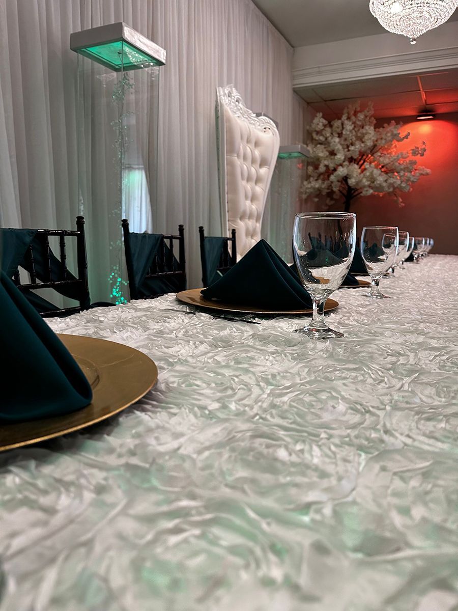 Long table set for an event; emerald napkins and gold chargers, white textured tablecloth.