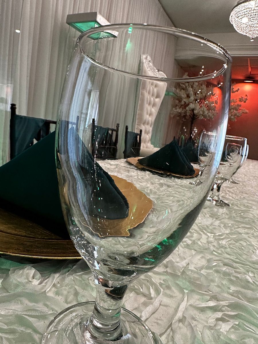 Clear wine glass reflecting a table setting with emerald green napkins, gold chargers, and a sparkly tablecloth.