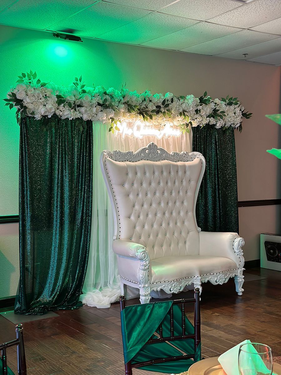 White throne-like chair against emerald green sequined curtains and floral garland. Bright lighting.