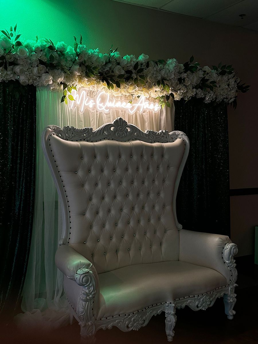 White, ornate loveseat in front of a backdrop with white flowers, lights, and green fabric.