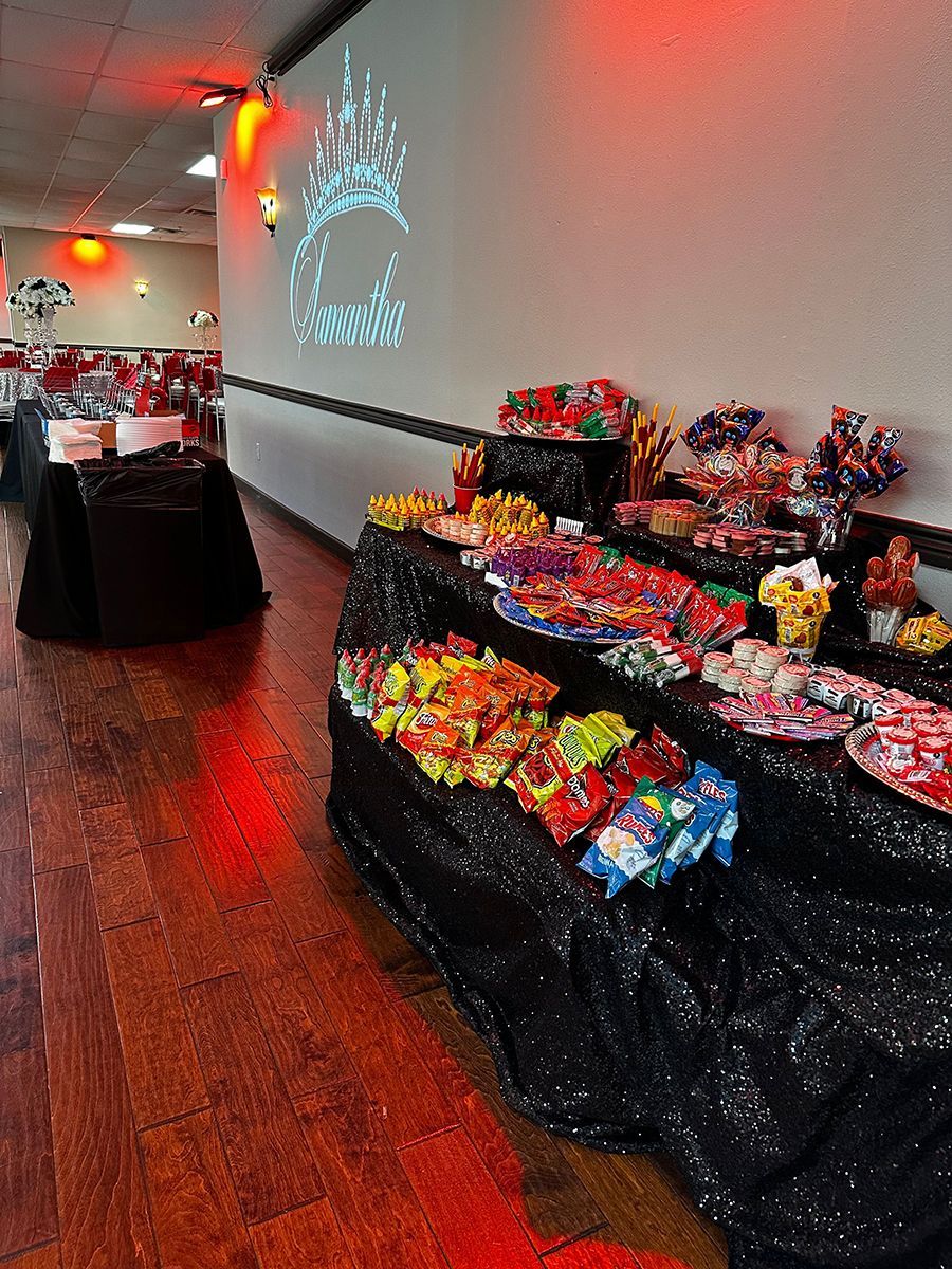 A brightly lit party room with tables of colorful candy and desserts. Red uplighting and a crown projection on the wall.
