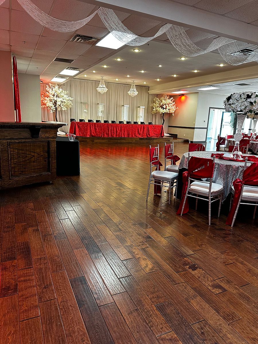 Empty banquet hall with wood floor, red table settings, and white floral arrangements.