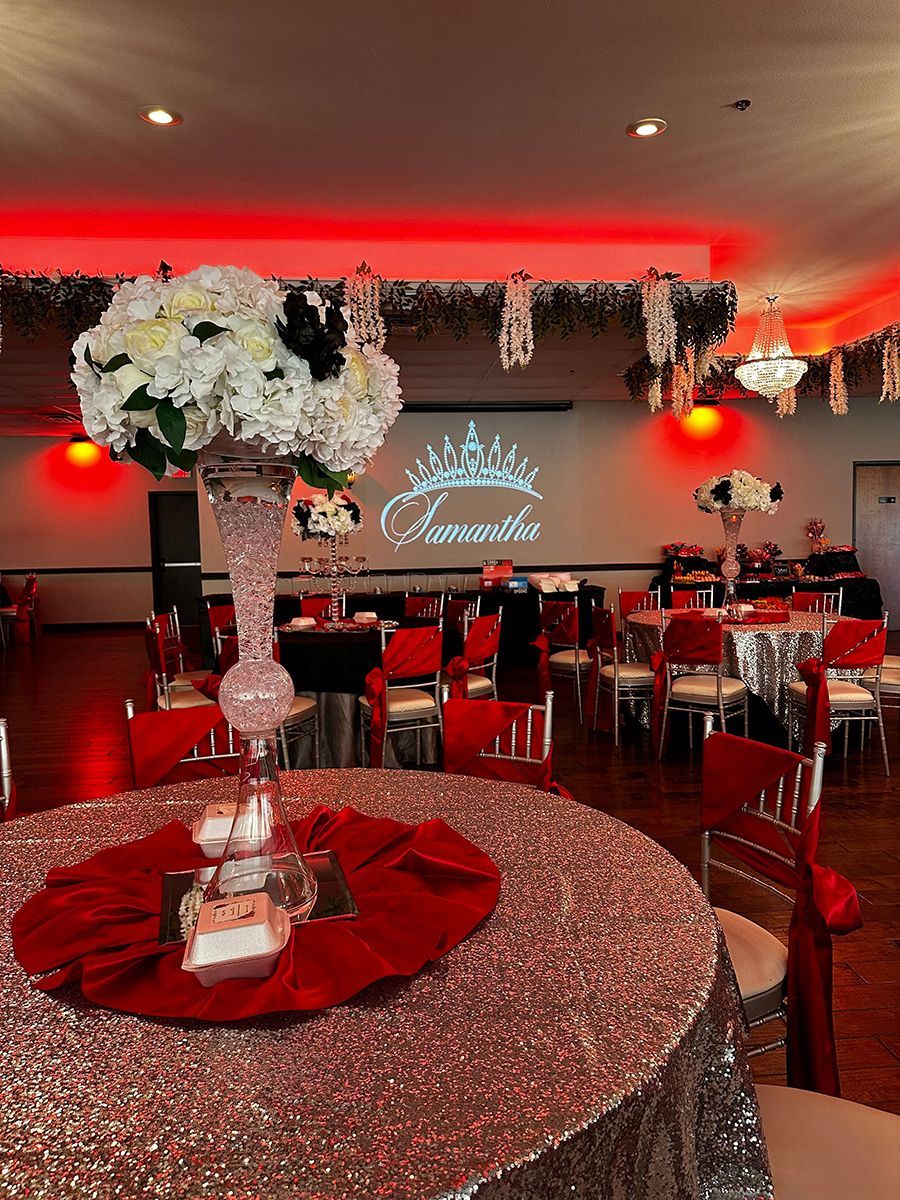 Elegant event hall with tables set for a party, featuring red and white floral centerpieces and red lighting.