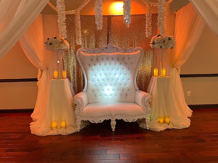 Elegant white throne chair against a gold sequined backdrop, decorated with flowers and candles.