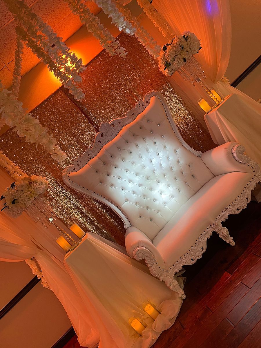 White ornate throne chair in a wedding stage setting, decorated with flowers and ambient lighting.