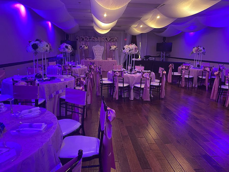 Event hall set for celebration, with pink tablecloths, floral centerpieces, and decorative chair sashes.