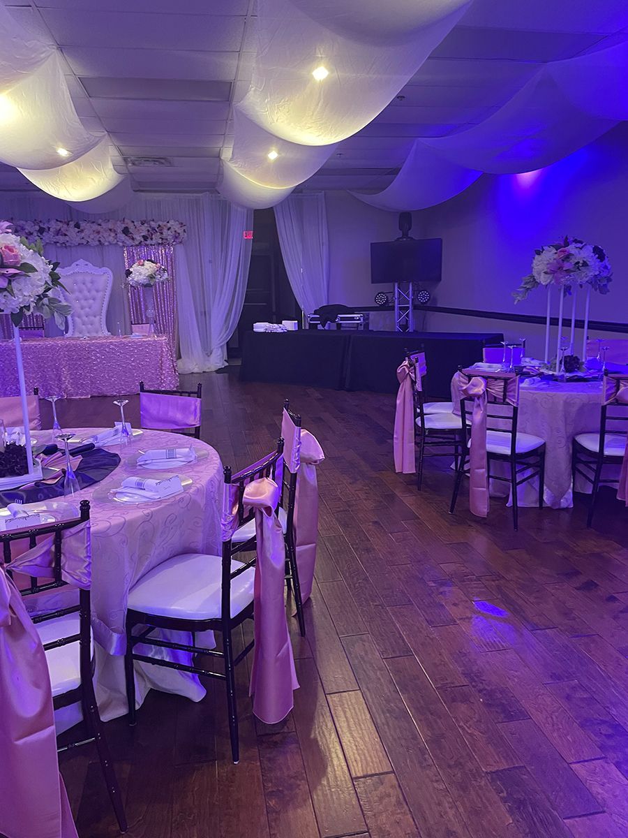 Elegant event space, decorated with pink, white, and purple accents. Tables set for guests, with draped ceiling.