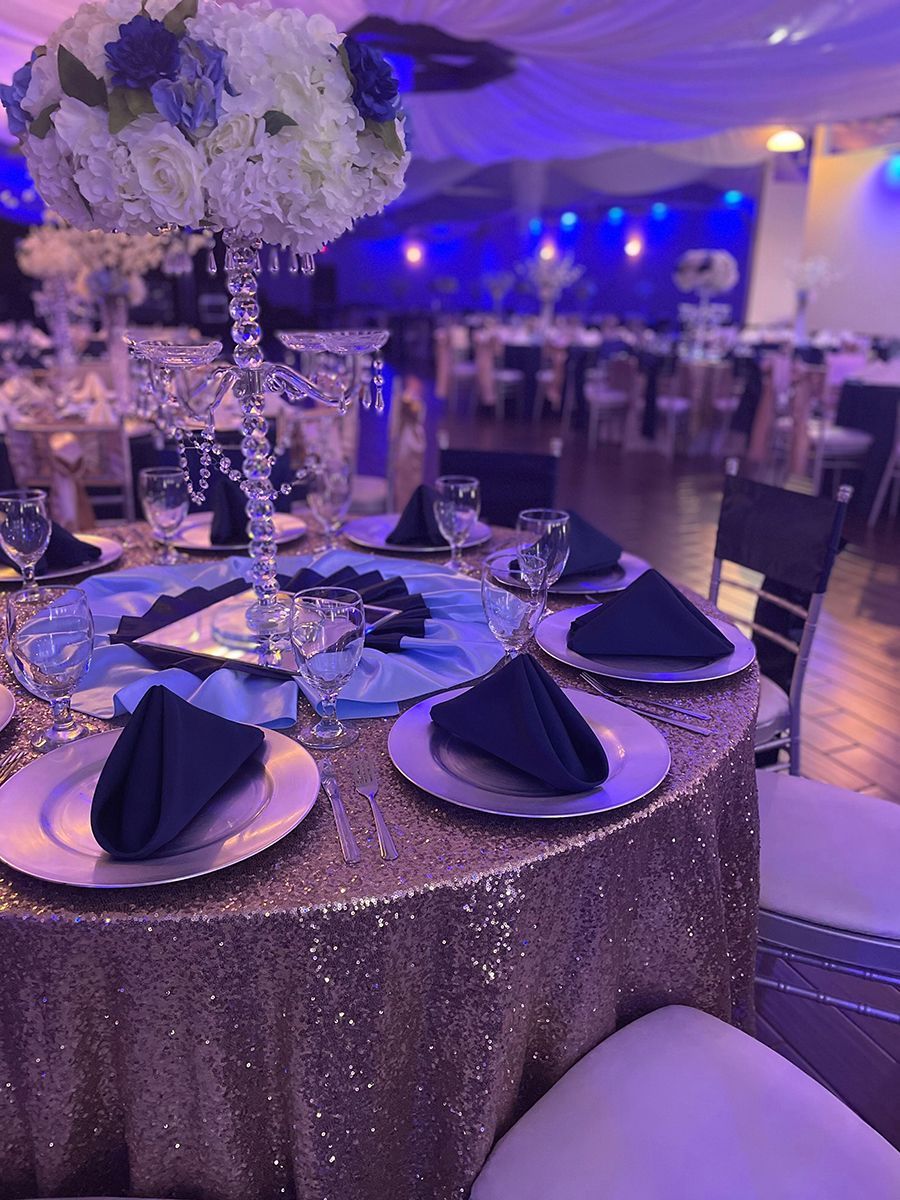 Table setting at a formal event with gold sequin tablecloth, floral centerpiece, and navy napkins.