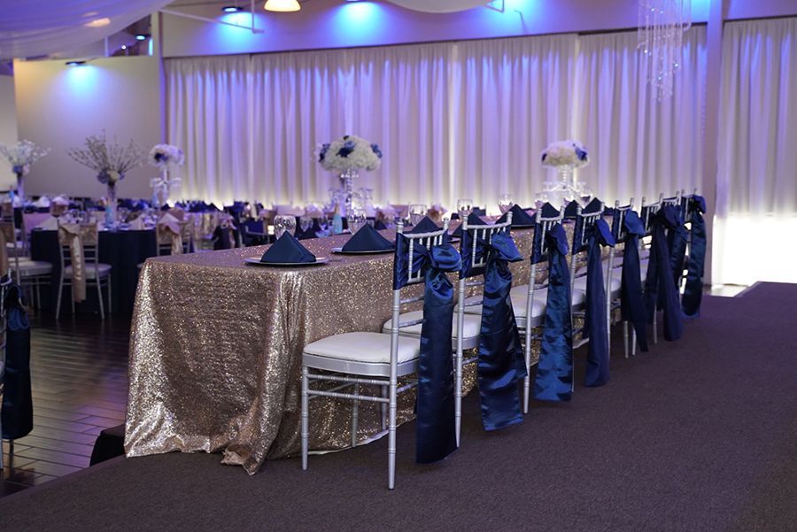 Banquet hall decorated for an event with a gold sequined head table, white chairs, and navy accents.