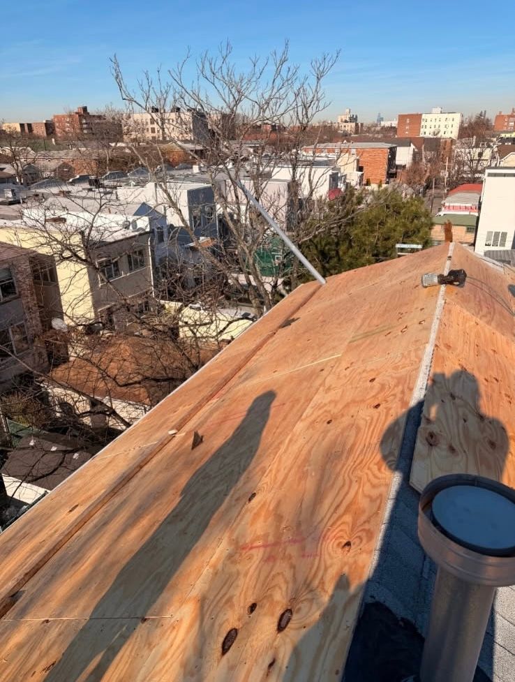 A view from a roof showing new plywood sheathing, a plumbing vent, and a residential neighborhood under a blue sky.