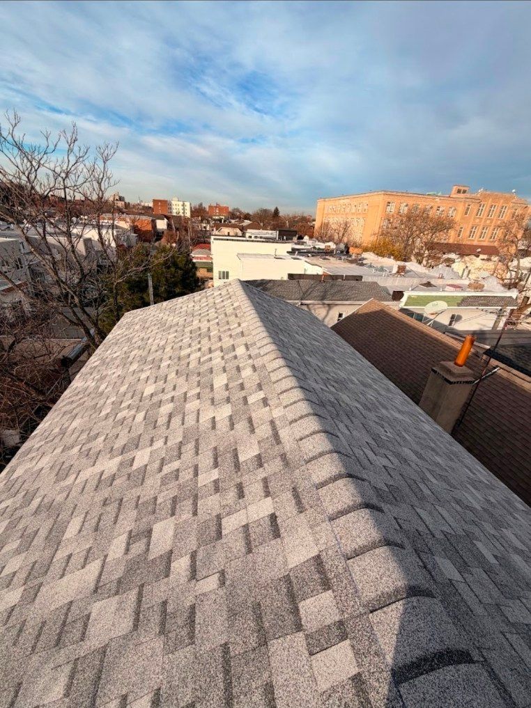 A high-angle view of a newly shingled gray roof under a blue sky, overlooking a suburban neighborhood.