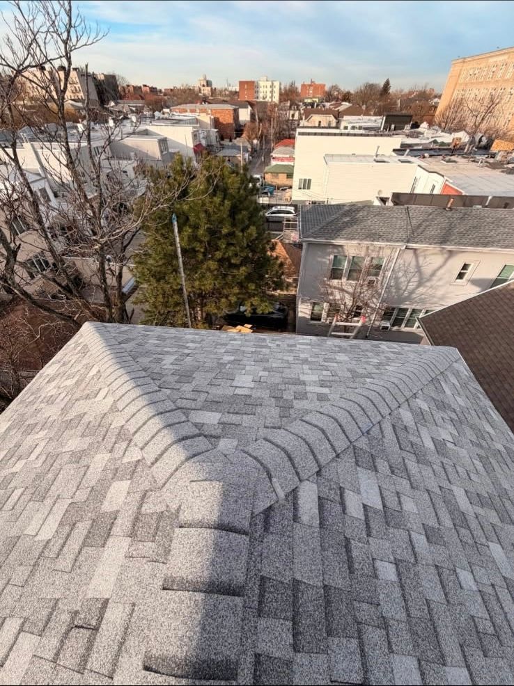 A high-angle view of a newly shingled gray roof with a three-way ridge cap, overlooking a suburban neighborhood.