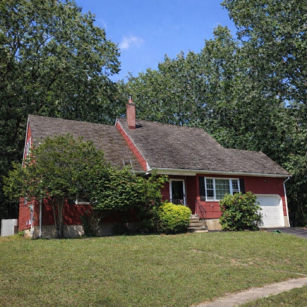 A red, single-story house with a grey shingled roof, set behind a green lawn and surrounded by mature leafy trees.