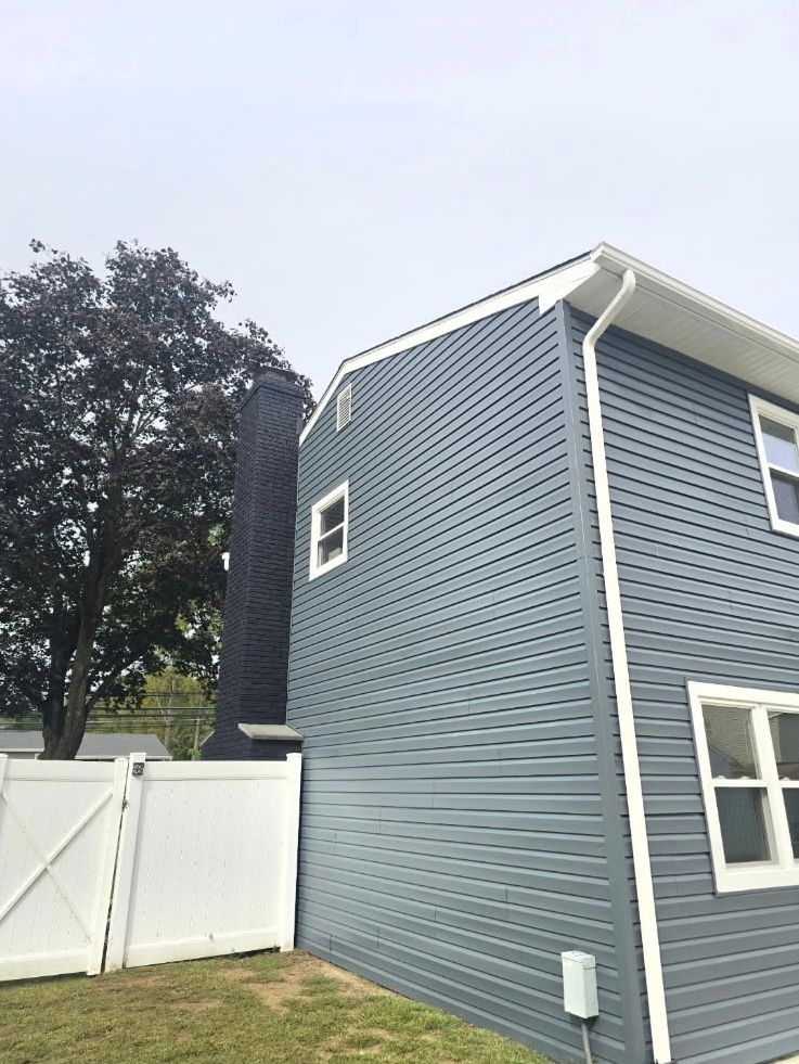 A blue-sided two-story house with a black chimney, white trim, and a white fence in the yard.