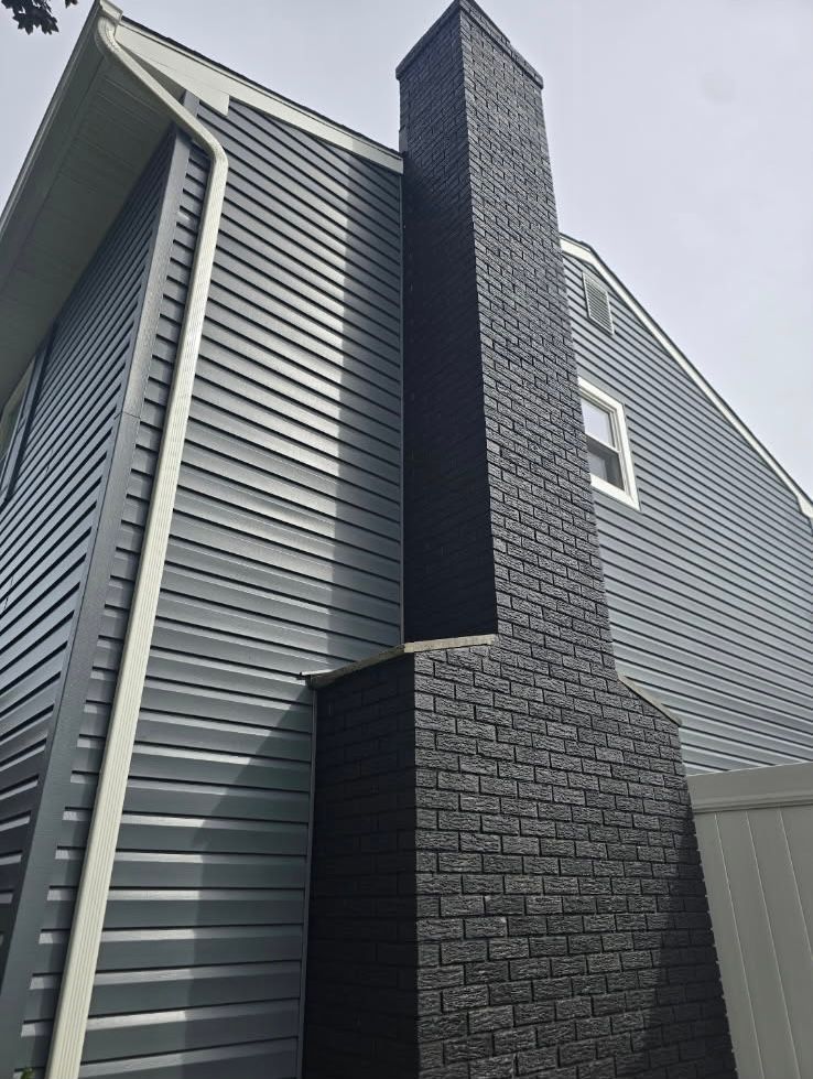 A tall, dark gray brick chimney extending up the side of a house with light gray horizontal vinyl siding.
