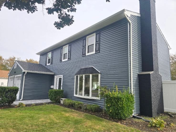 A two-story blue-sided house with a large bay window, a front garage, and a black brick chimney on the side.
