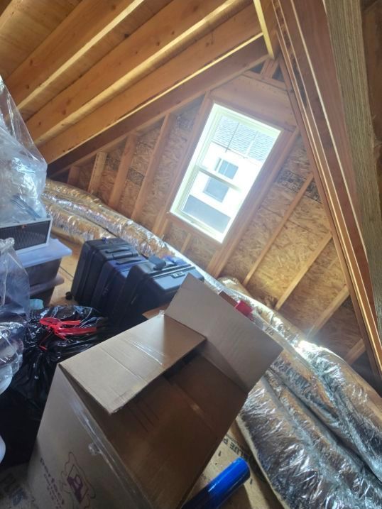 An attic space with exposed wood beams, a small window, storage boxes, and flexible ductwork on the floor.