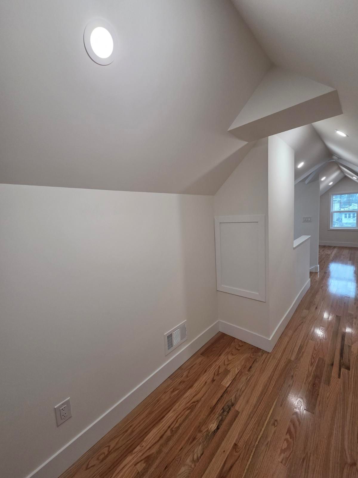 An attic hallway with hardwood floors, white slanted walls, a ceiling light, and a window in the distance.