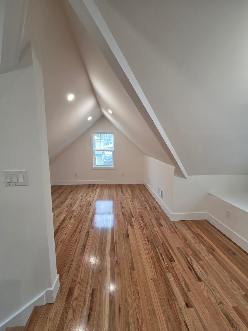 An empty attic room with vaulted white ceilings, light wood floors, and a single window at the far end.
