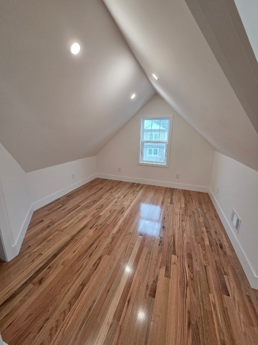 An empty attic room with beige walls, sloped ceilings, recessed lighting, and polished hardwood floors with a window.