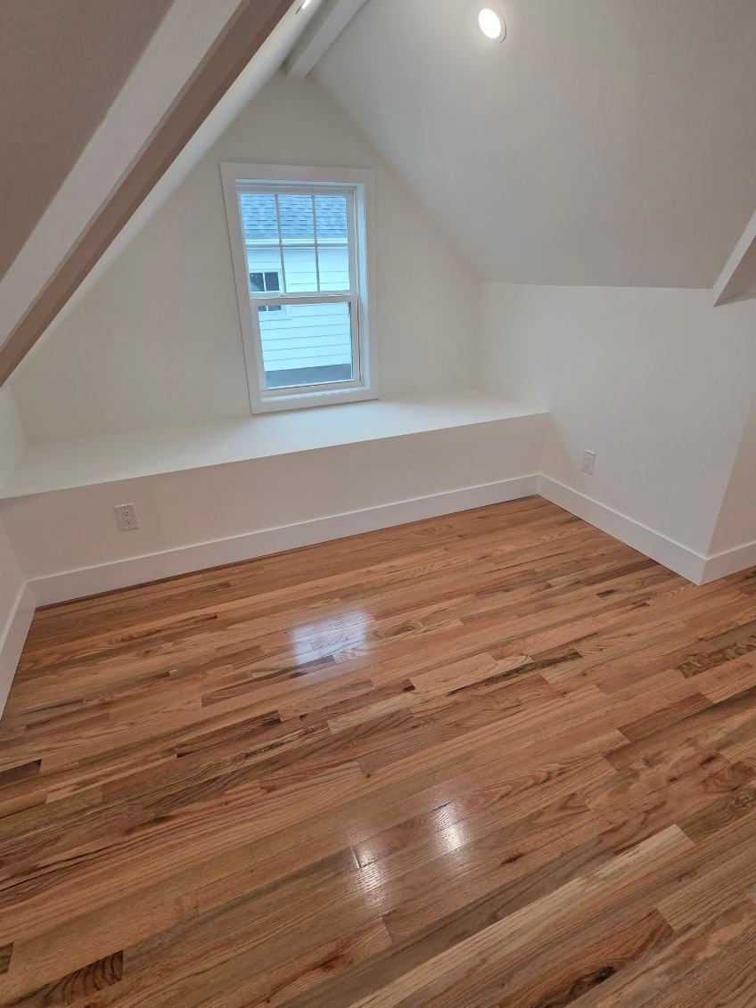 An empty attic room with white slanted walls, hardwood floors, and a built-in window bench seat.