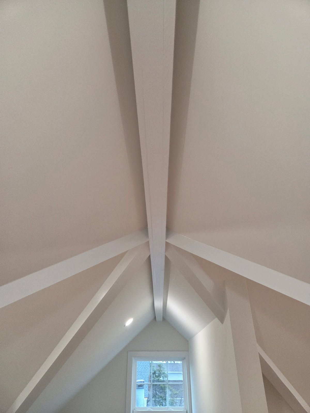 A low-angle view of a white, vaulted attic ceiling featuring exposed structural beams and a window at the far end.