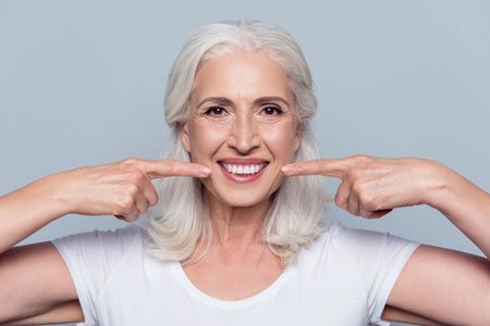 An older woman is smiling and pointing at her teeth.