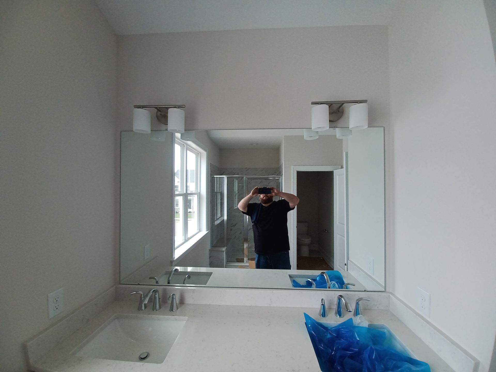 Bathroom mirror reflecting a person taking a selfie, with sink, counter, and blue toiletries in the foreground