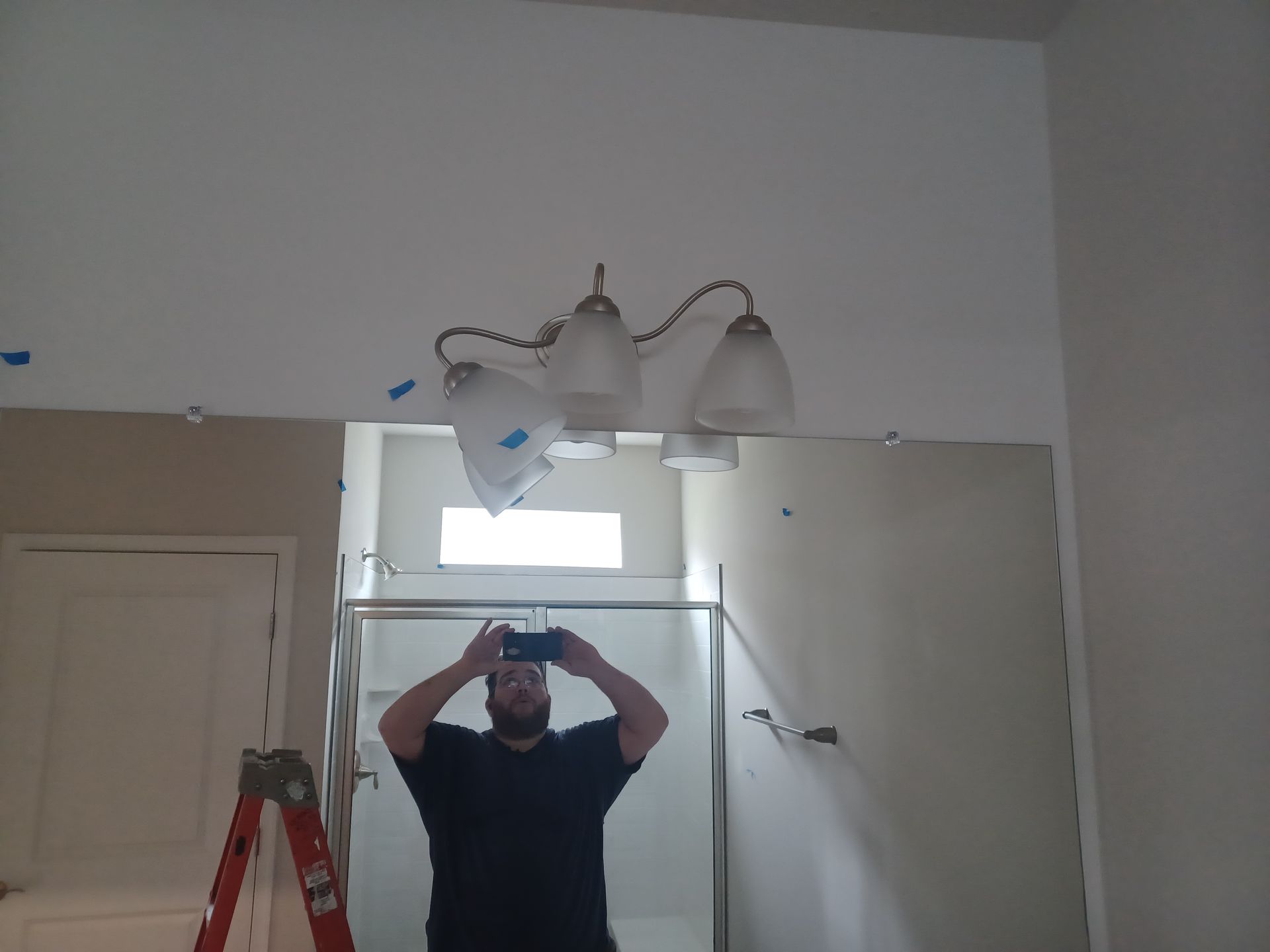 Person taking a mirror selfie in a bathroom with a red ladder and wall lights above the mirror