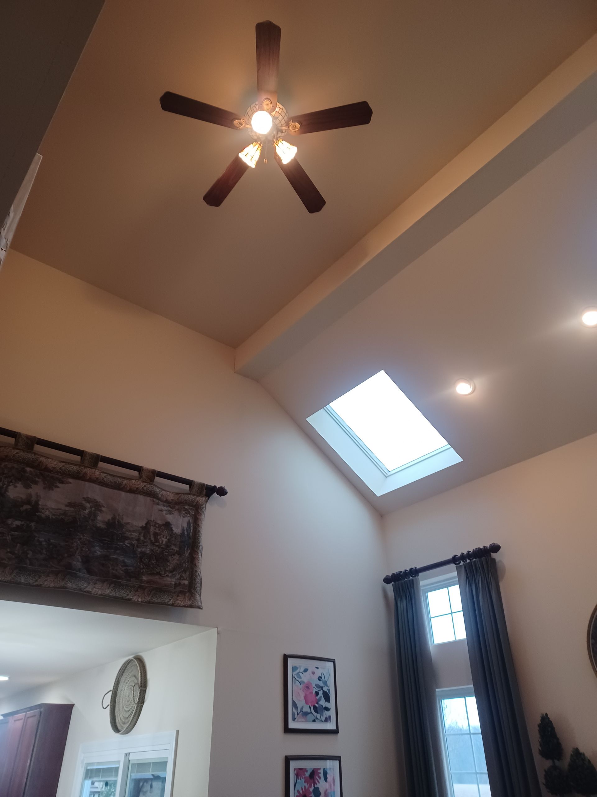 Living room ceiling with a lit ceiling fan and skylight, beige walls, curtains, and framed wall art.
