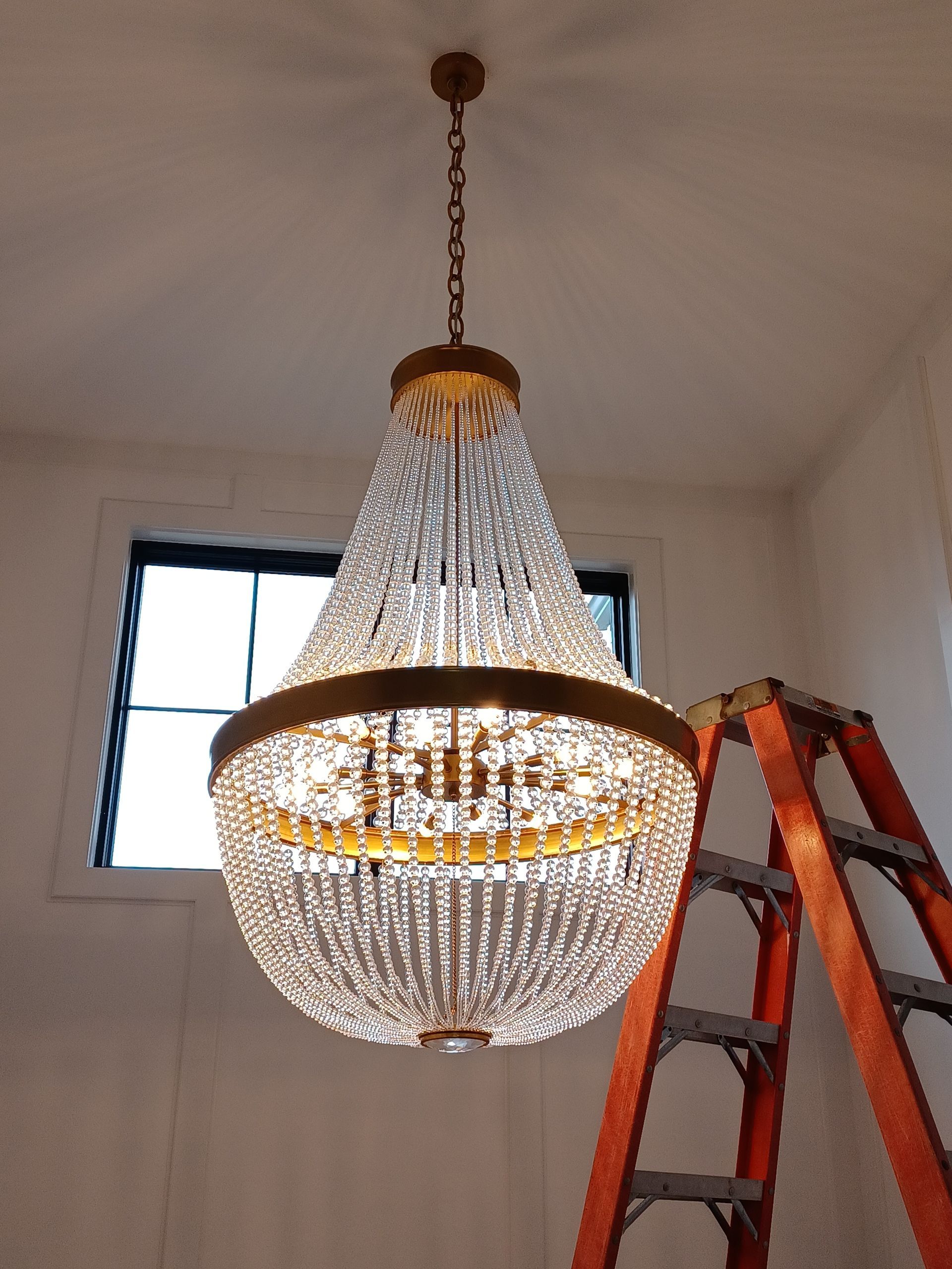 Crystal chandelier hanging in a room, with a red ladder beside it and a window behind.