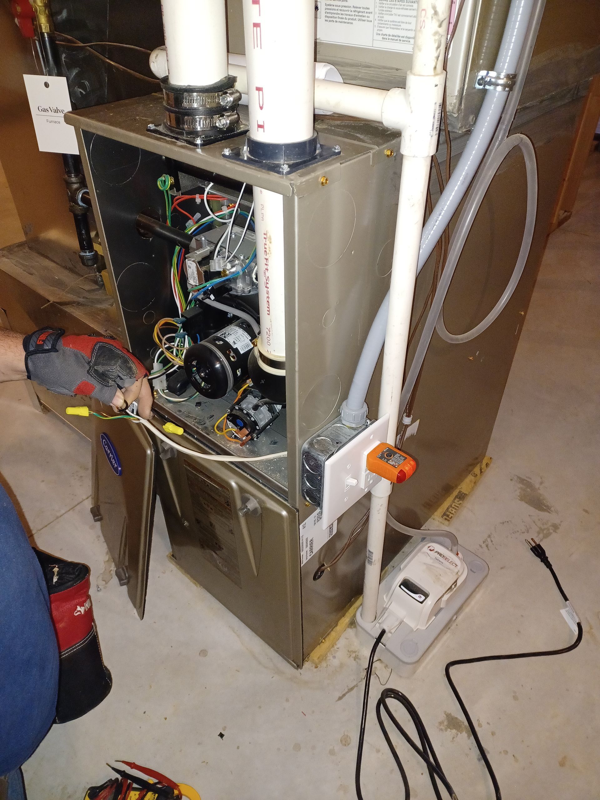 Open furnace cabinet with exposed wiring and pipes on a basement floor, tools nearby