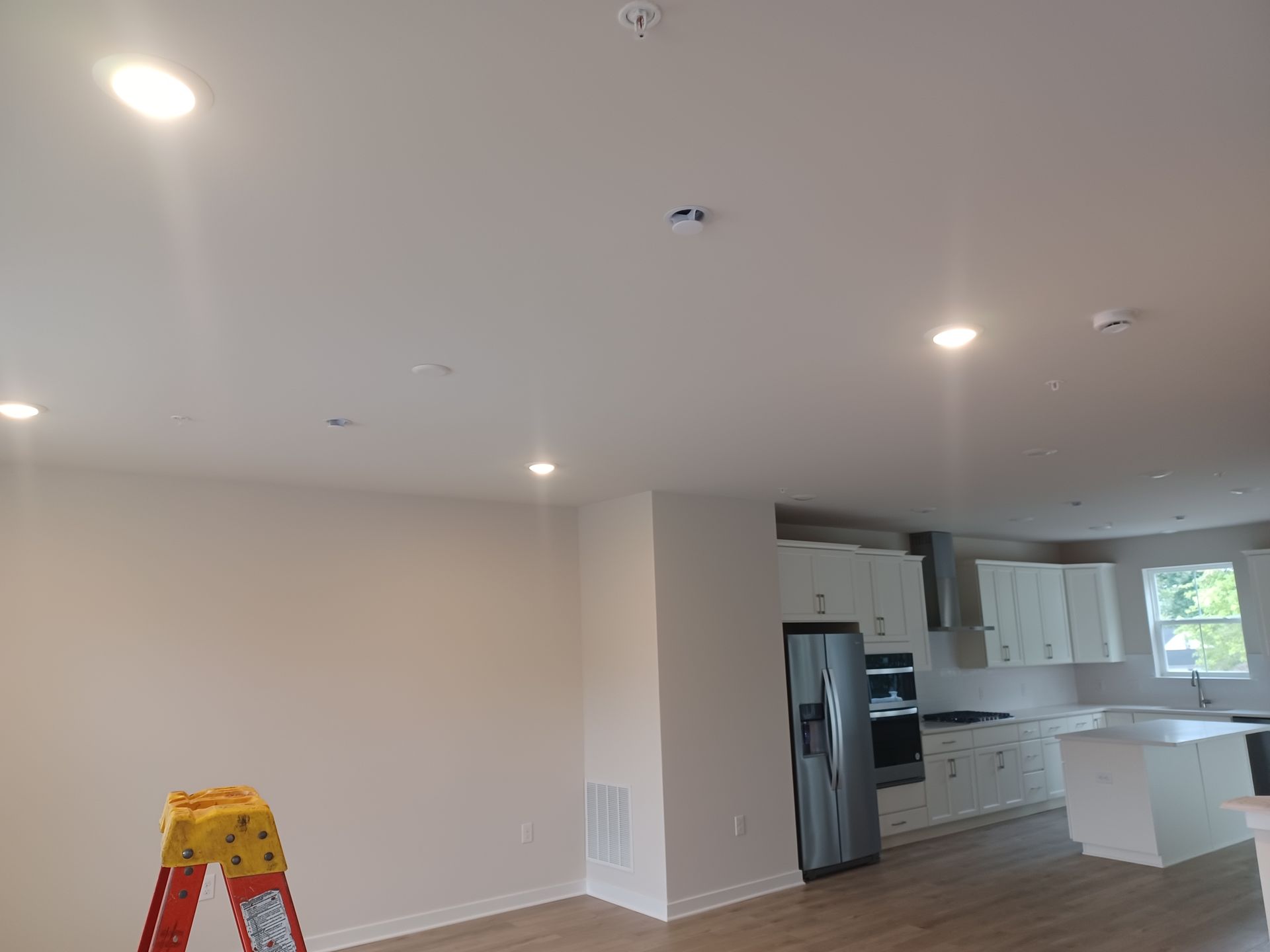 Empty living room and kitchen with white walls, recessed lights, and a step ladder in the foreground.