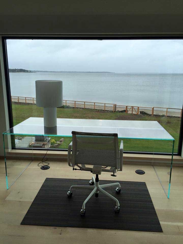 Glass desk and chair with ocean view through a large window. A lamp sits on the desk, with a rug below the chair.