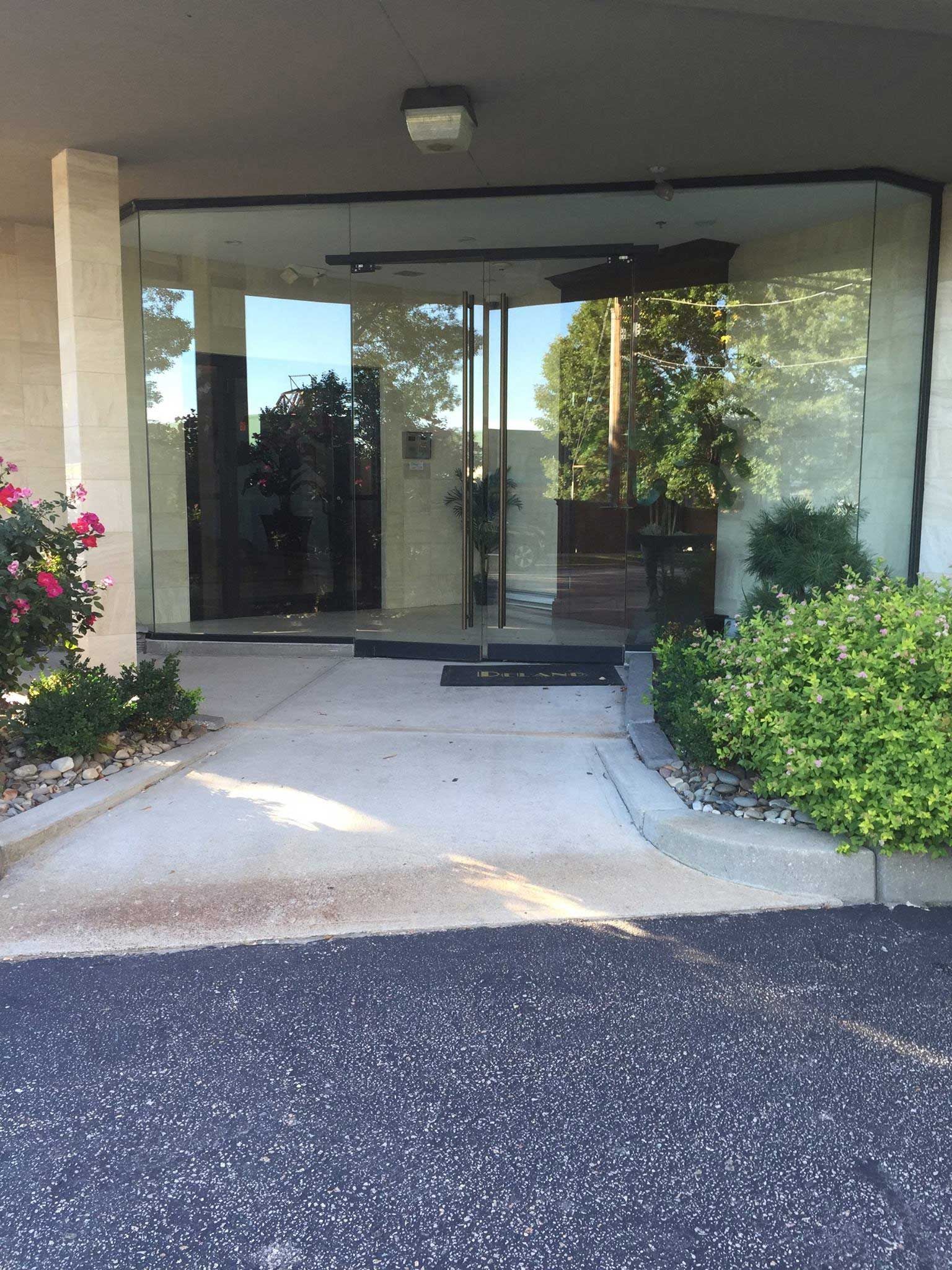 Glass entrance to a building. Concrete path leads to clear glass doors and windows. Plants and rose bushes frame the entrance.
