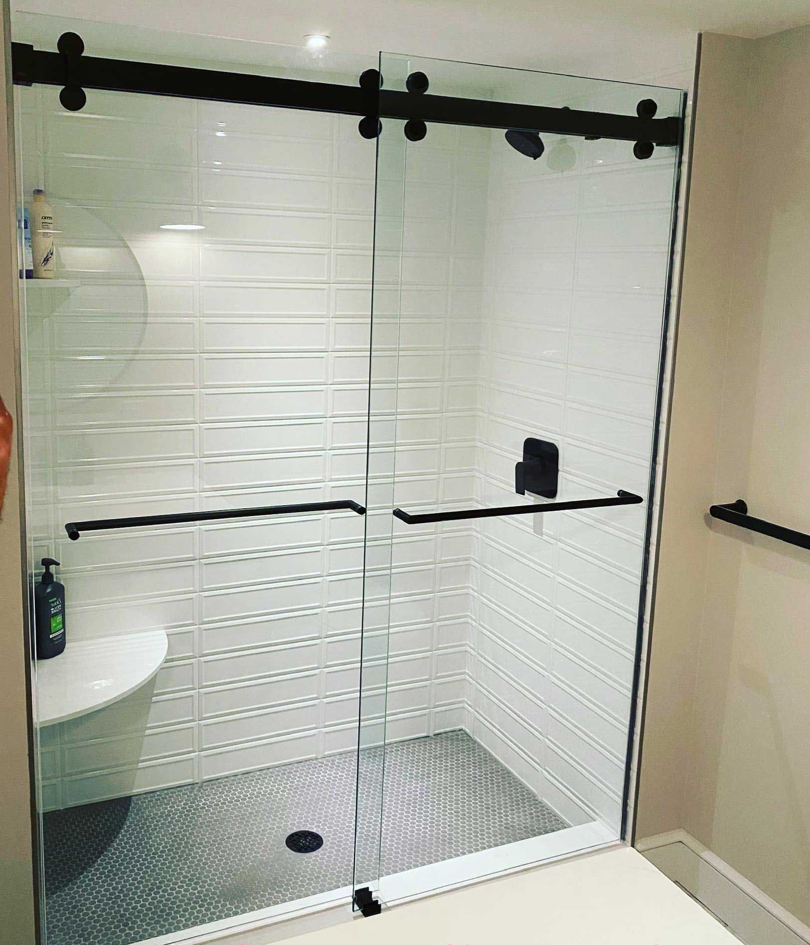 Shower with sliding glass doors, black hardware, white tile walls, and gray pebble floor.