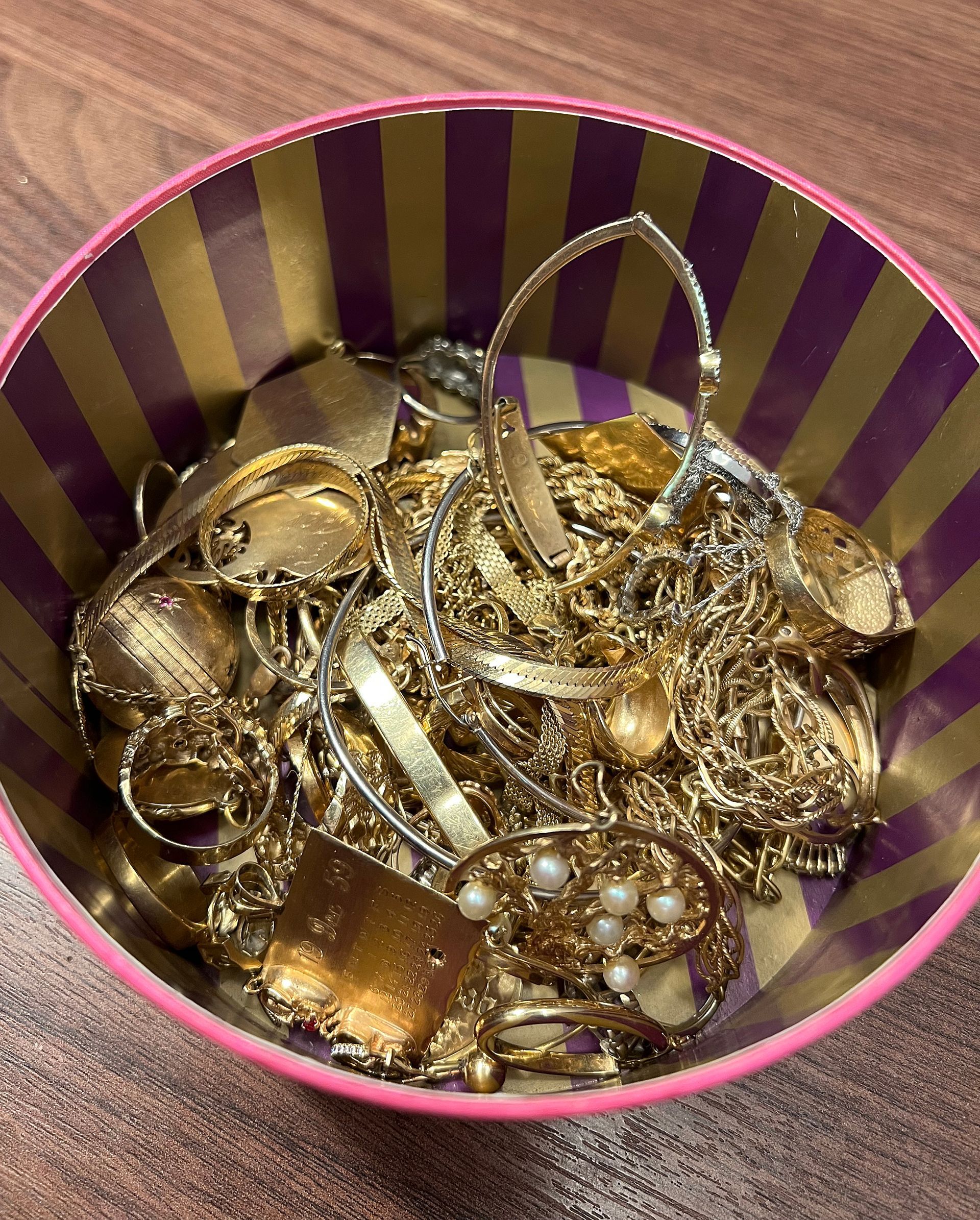 A bowl filled with gold jewelry is on a wooden table.