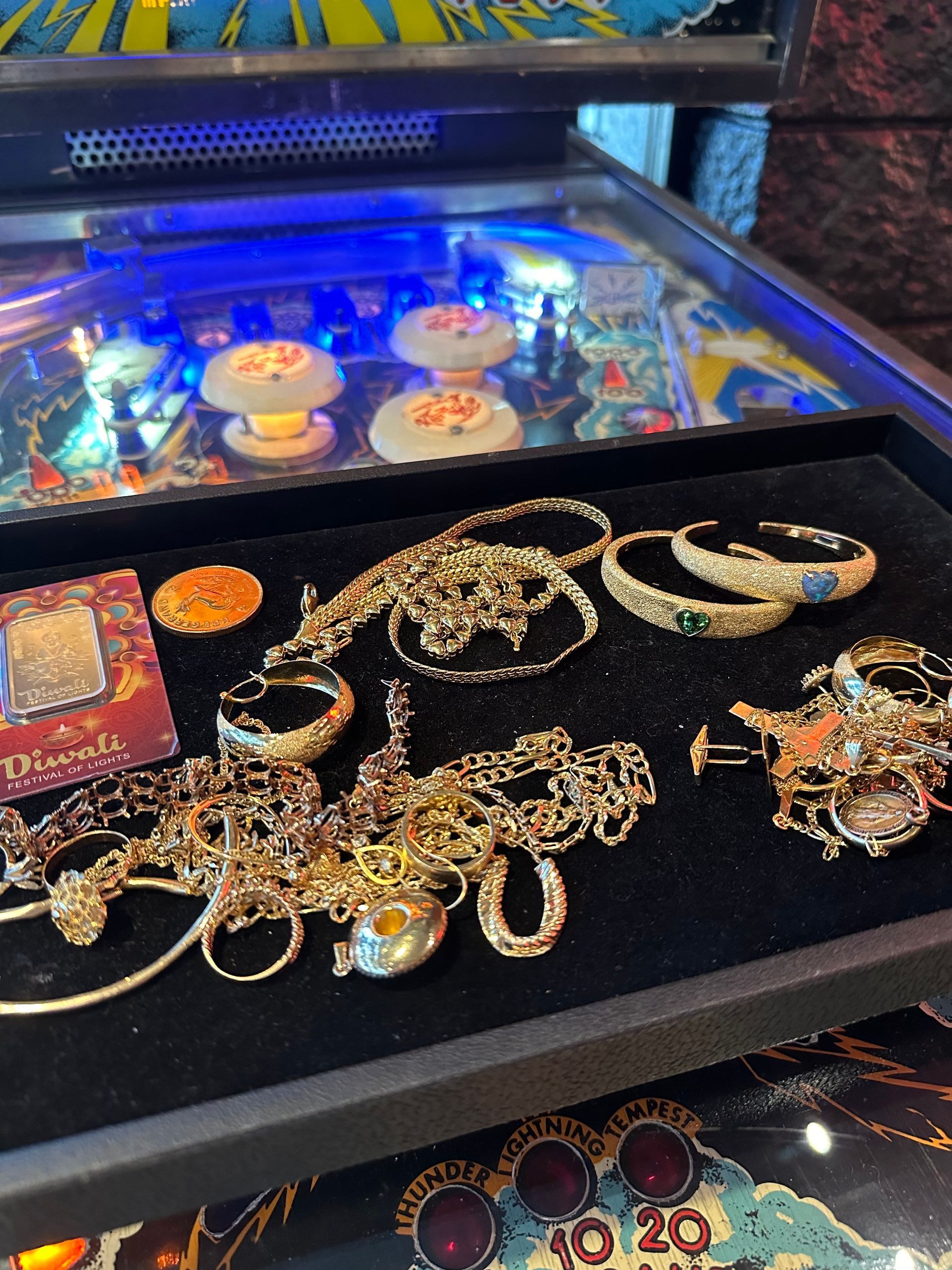 Black velvet tray displays gold jewelry near a pinball machine's playfield.