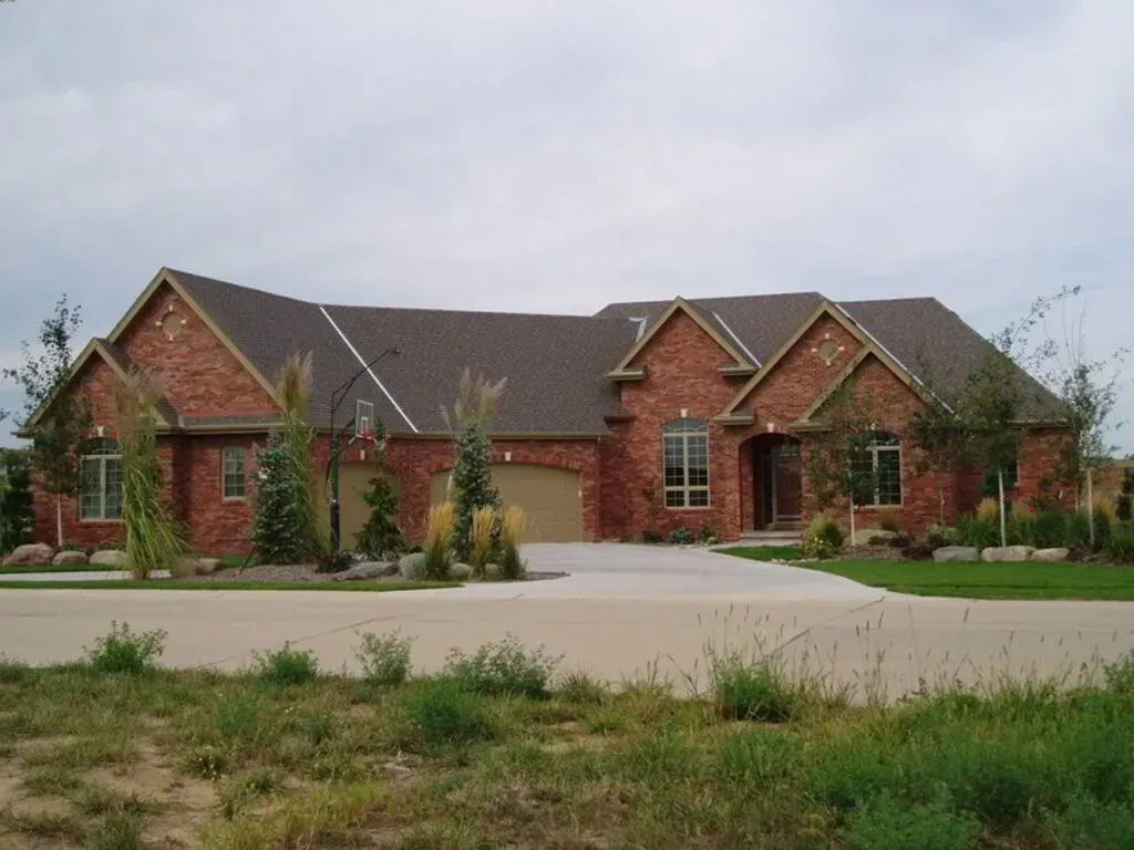 A one-story red brick house with a multi-gabled roof and a three-car garage, surrounded by landscaped trees and shrubs.