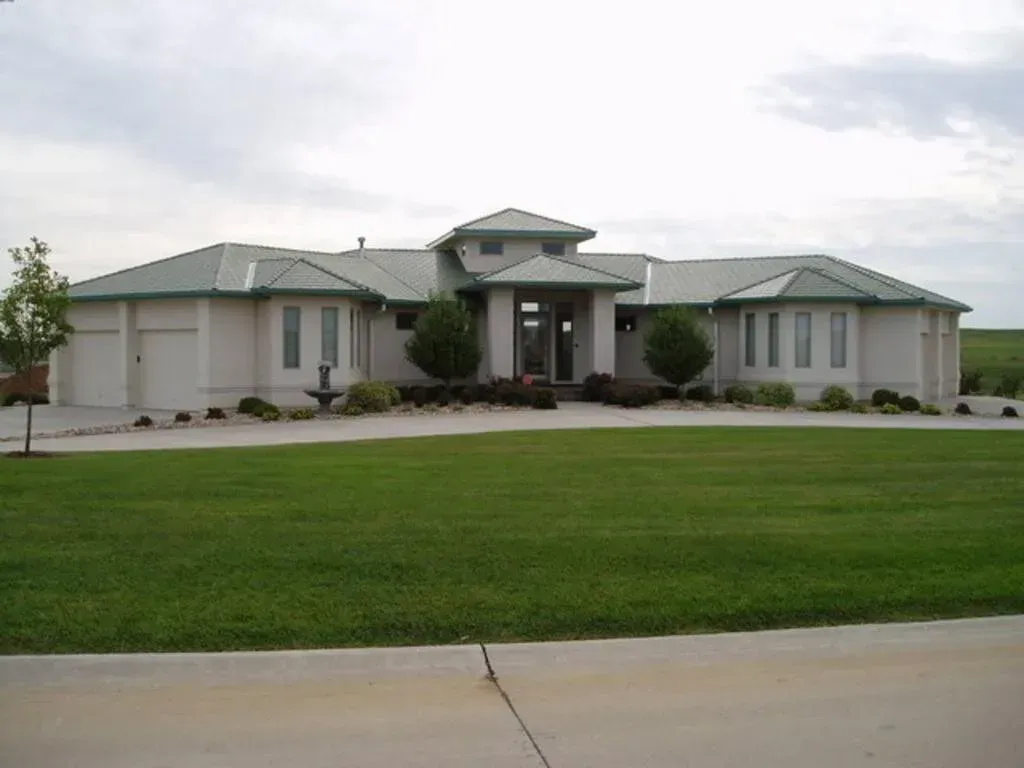 A light-colored, single-story modern house with a green roof, a central entryway, and a large front lawn under a cloudy sky.