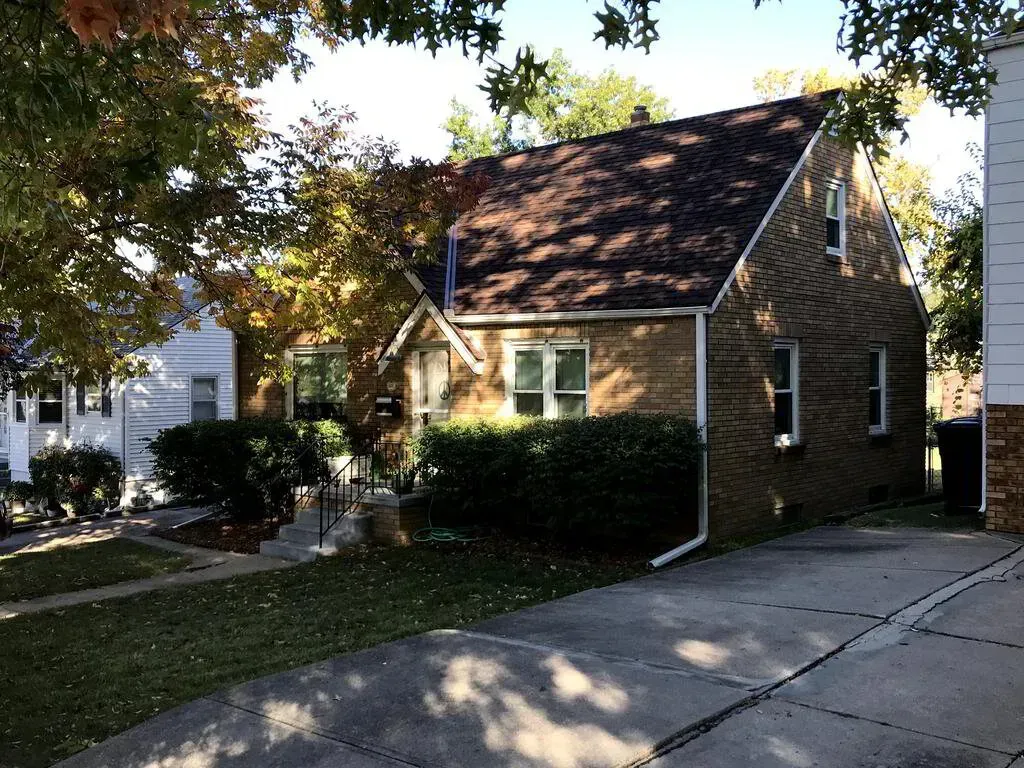A brown brick house with a dark gabled roof and a concrete driveway on a sunny day with trees in the foreground.