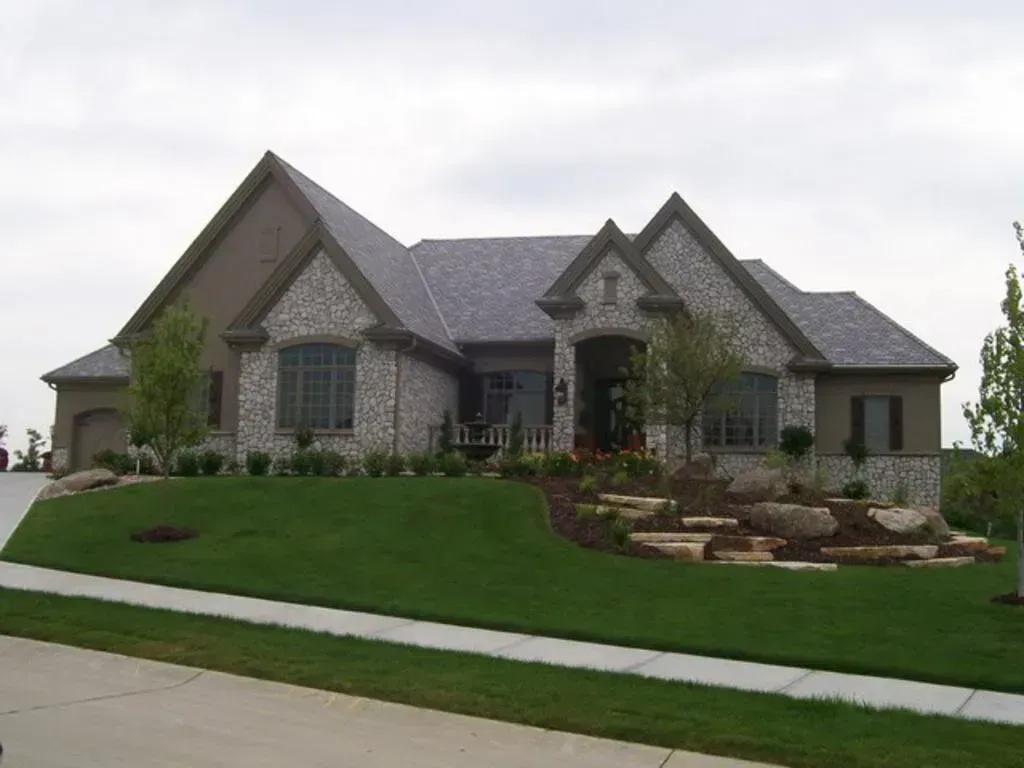 A multi-gabled house with grey stone and brown siding, a manicured lawn, and stone landscaping features on a cloudy day.